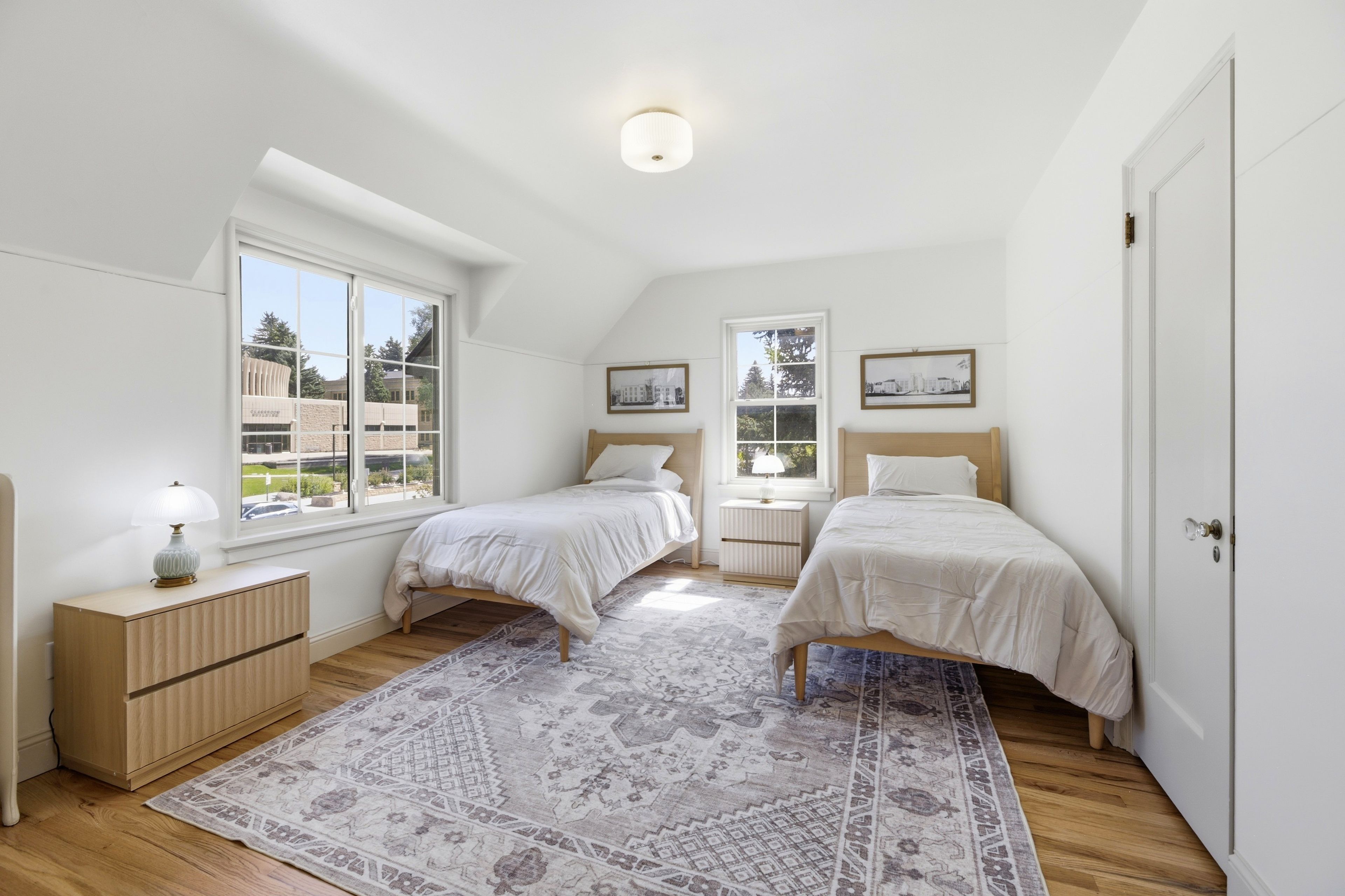 Een slaapkamer met twee eenpersoonsbedden, een nachtkastje met een lamp, een dressoir, een kamerkleed en twee ramen met uitzicht op de stad.