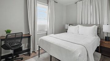 A bedroom with a bed, a desk with a chair, a bedside table with a lamp, and a window with curtains.