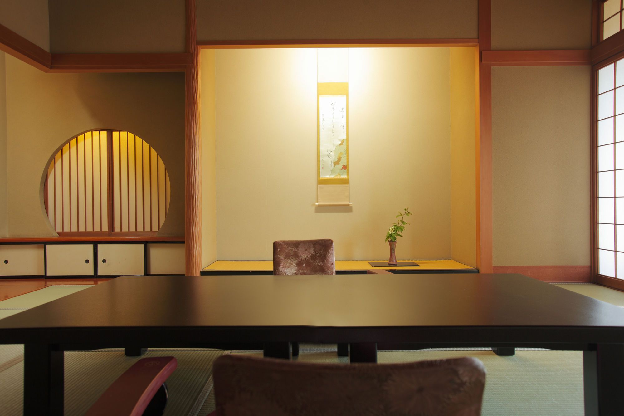 A traditional Japanese room with tatami flooring, a low table, a cushioned chair, a small table with a plant, and a sliding door with a decorative panel.