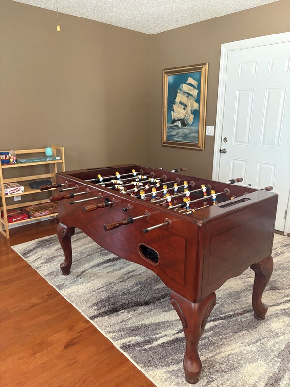 A wooden foosball table in a room with a striped rug, a bookshelf with board games, and a framed picture on the wall.