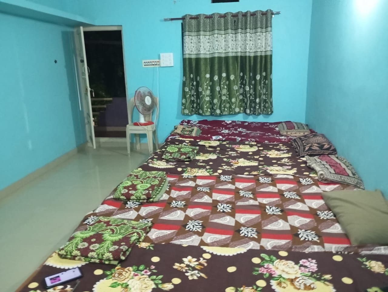 Una habitación con una cama cubierta con ropa de cama estampada, un ventilador de pie, una ventana con cortinas y una puerta que da a otra habitación.