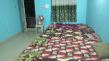 Una habitación con una cama cubierta con ropa de cama estampada, un ventilador de pie, una ventana con cortinas y una puerta que da a otra habitación.