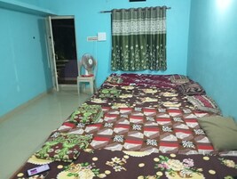 A room with a bed covered in patterned bedding, a standing fan, a window with curtains, and a door leading to another room.