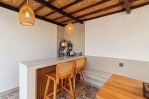 Una cocina con mostrador blanco, dos sillas de mimbre, una mesa de madera y techo tejido.