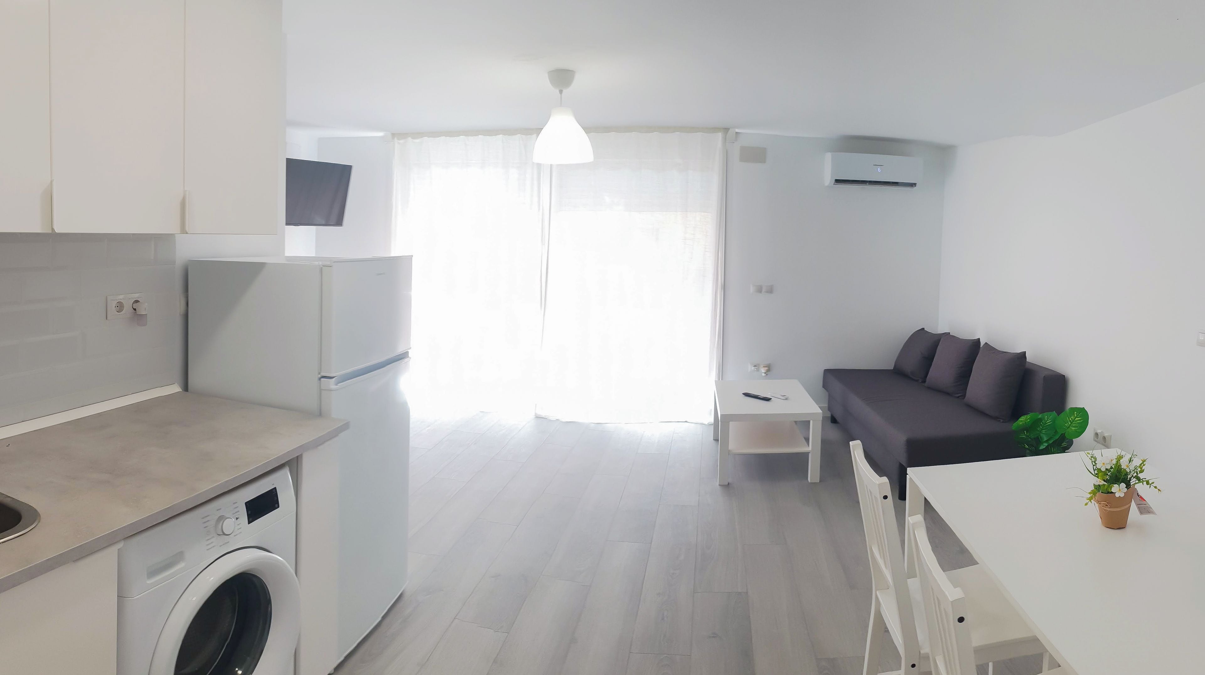 A modern kitchen with a washing machine, a dining area with a table and chairs, a gray sofa, and a large window with sheer curtains.