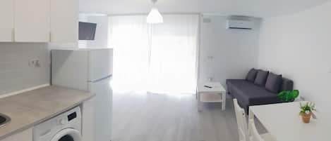 A modern kitchen with a washing machine, a dining area with a table and chairs, a gray sofa, and a large window with sheer curtains.