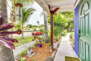 A covered porch with a statue, a blue door, and a garden with various plants and a fence.