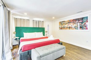 Habitación de hotel con cama, mesita de noche, lámpara, ventana con cortinas, cabecera verde, reposapiés gris y un cuadro de un paisaje urbano colorido en la pared.