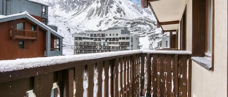 A snow-covered balcony with a mountain view, wooden railings, and a building with multiple windows.