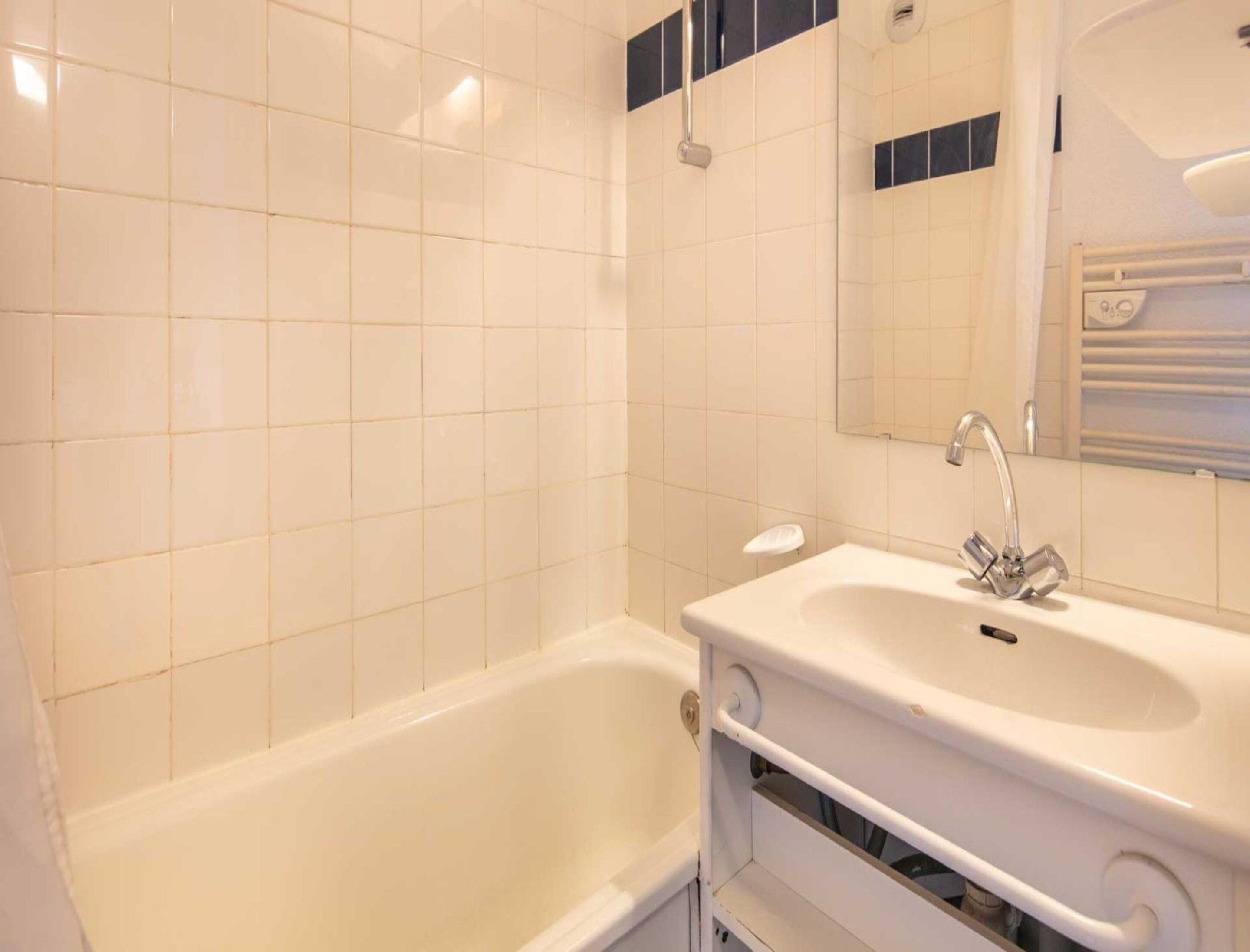 Ein Badezimmer mit weißen Fliesen, einer Badewanne, einem Waschbecken inklusive Spiegel und einem Handtuchhalter.