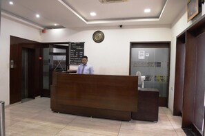 A reception desk in a hotel lobby with a digital display board, a clock, and a door to the right. - OYO Rooms Sampatrao Colony Alkapuri (Vadodara)