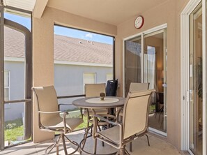 Eine überdachte Terrasse im Freien mit Tisch und Stühlen, einer Uhr an der Wand und Blick auf ein Gebäude mit Satteldach.
