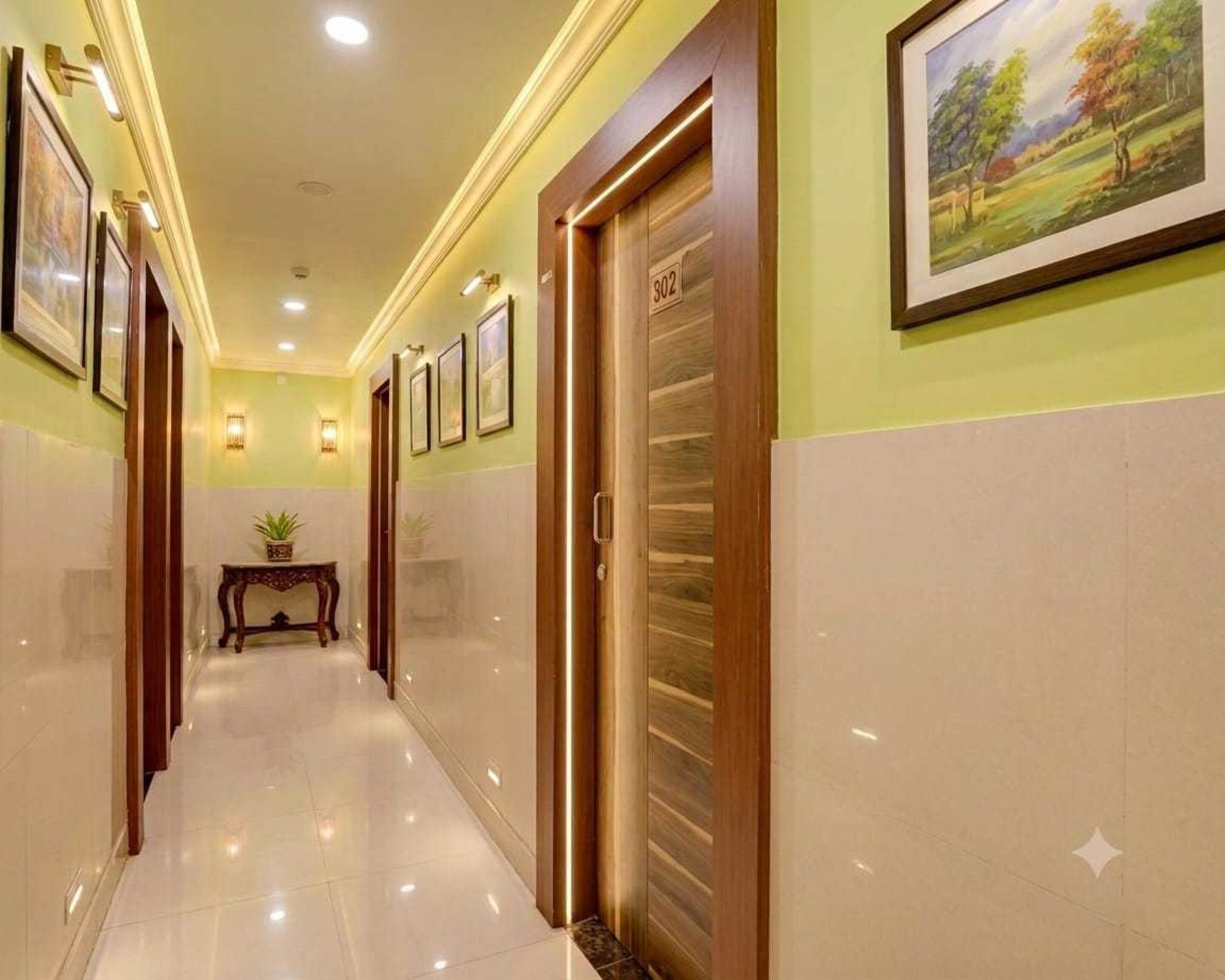 A hotel hallway with a polished floor, framed pictures on the walls, a wooden door with a number, and a small table with a plant.