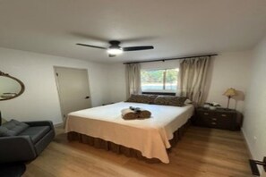 Quarto com cama, ventilador de teto, janela com cortinas, mesa de cabeceira e criado-mudo.