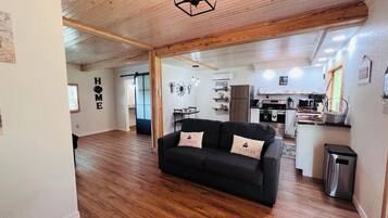 Un salon avec un canapé noir, un parquet en bois et une cuisine à l’arrière-plan.