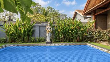 Una piscina con una estatua en el centro, rodeada de exuberante vegetación y un edificio con techo de tejas.