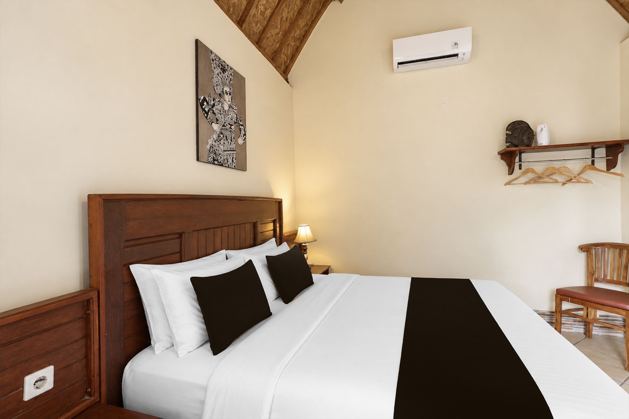 Una habitación de hotel con cabecera de madera, cama con ropa de cama blanca y negra, aire acondicionado de pared, lámpara de noche y una silla de madera.