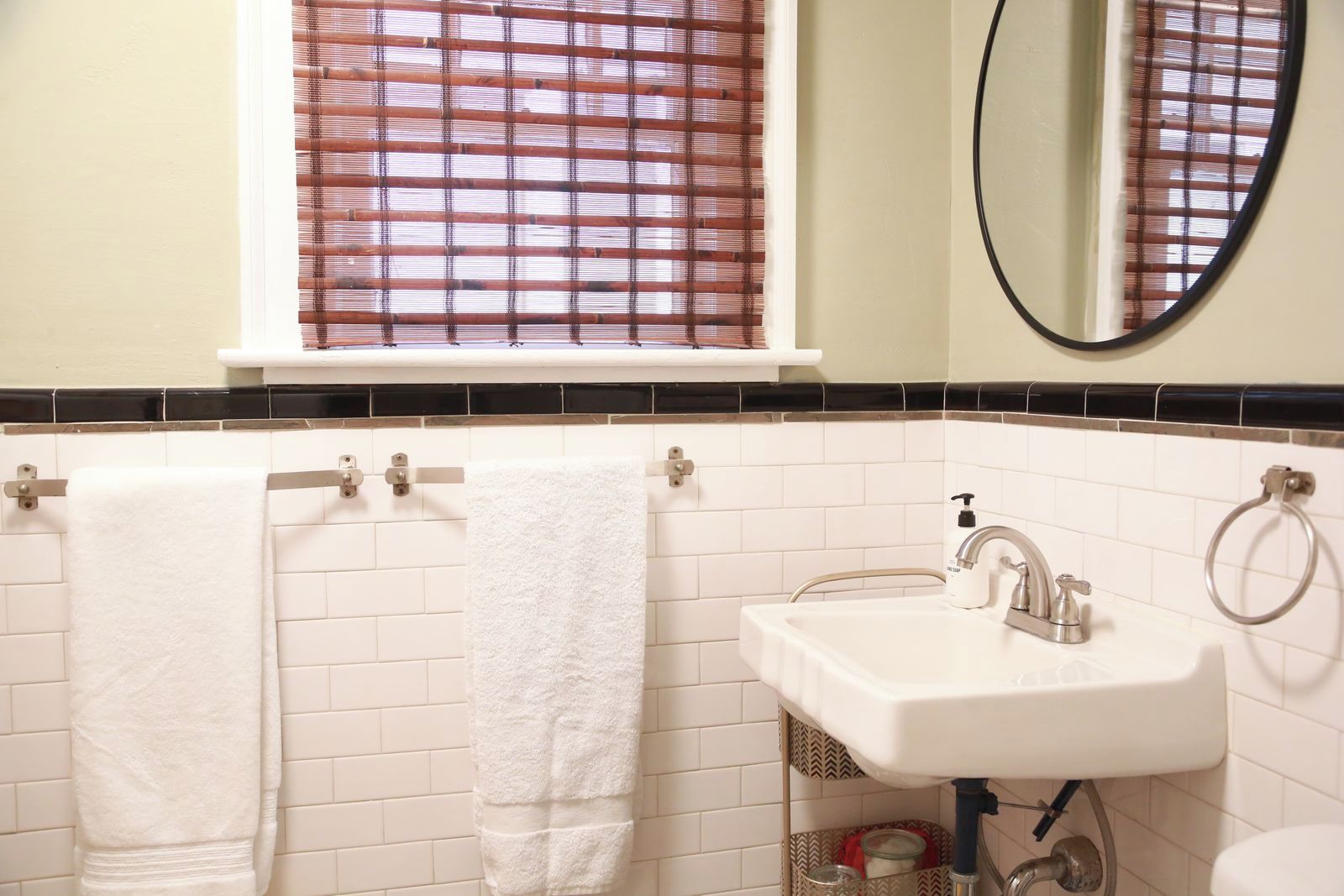 A bathroom with two white towels hanging on towel bars, a white sink with a mirror above it, and a window with a brown striped blind.