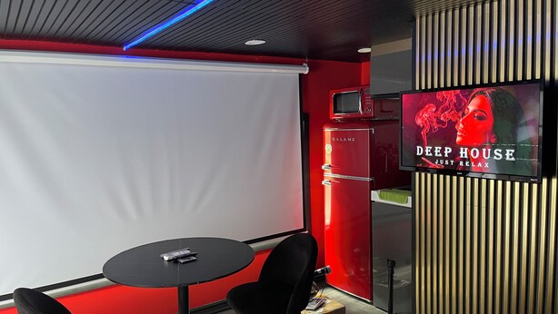 A room with a projector screen, a red wall, a refrigerator, a television displaying 'DEEP HOUSE', and a small table with chairs. - 1 Red Room private bathroom
In Luxury KIA   A1 (Richmond Hill)