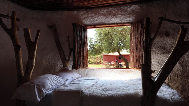 A rustic room with a bed, a window with curtains, and a view of the outside. - Maasai Heritage Museum (Kajiado County)