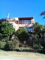 Un edificio de dos pisos con un balcón, rodeado de vegetación y una cerca.