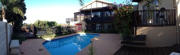 Una piscina rodeada por una terraza de madera, un área cercada y un edificio con balcones.