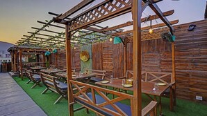 Área de estar ao ar livre com pérgola de madeira, mesas e cadeiras, cercada por um muro de madeira.