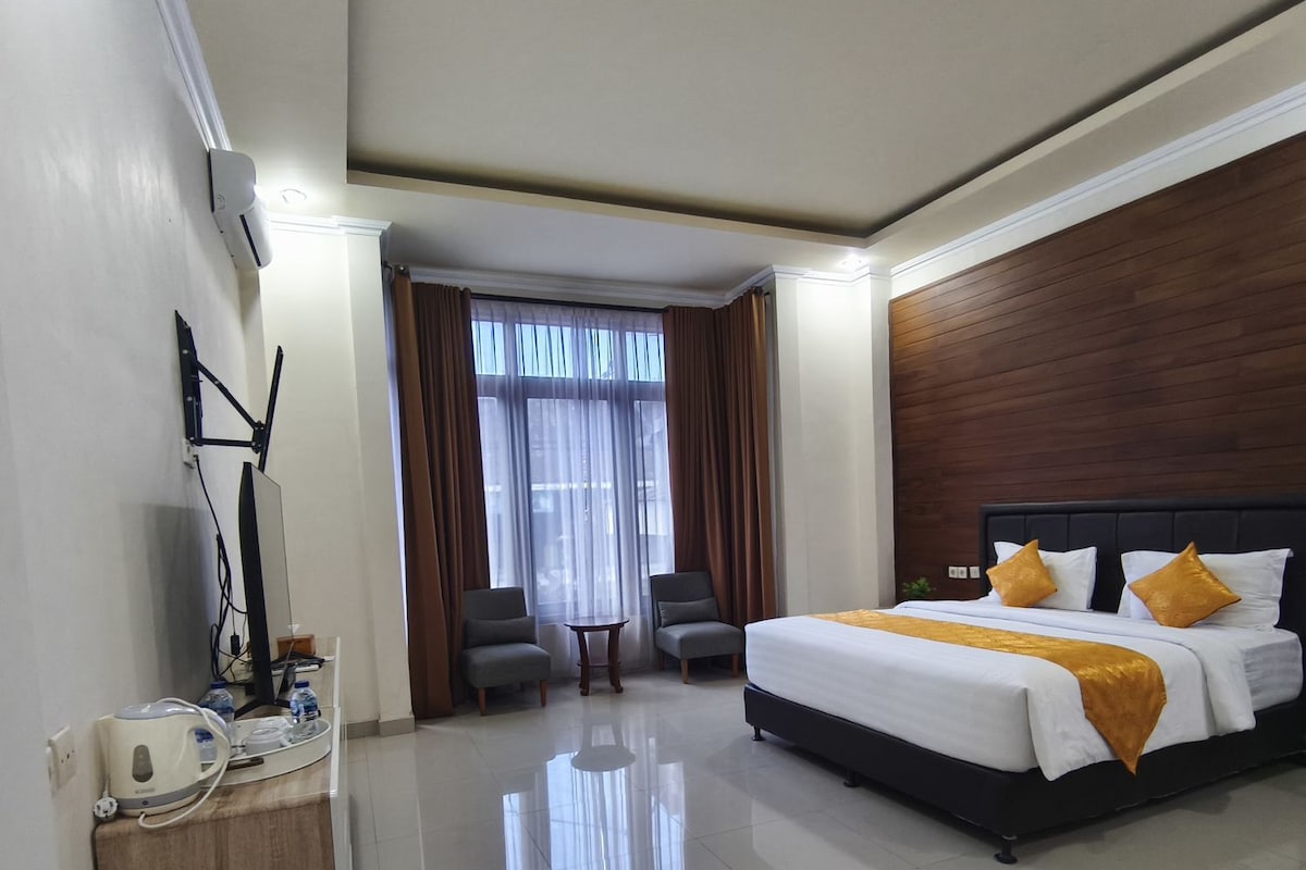 Habitación de hotel con una cama grande, un escritorio con tetera, una silla y un ventanal con cortinas.