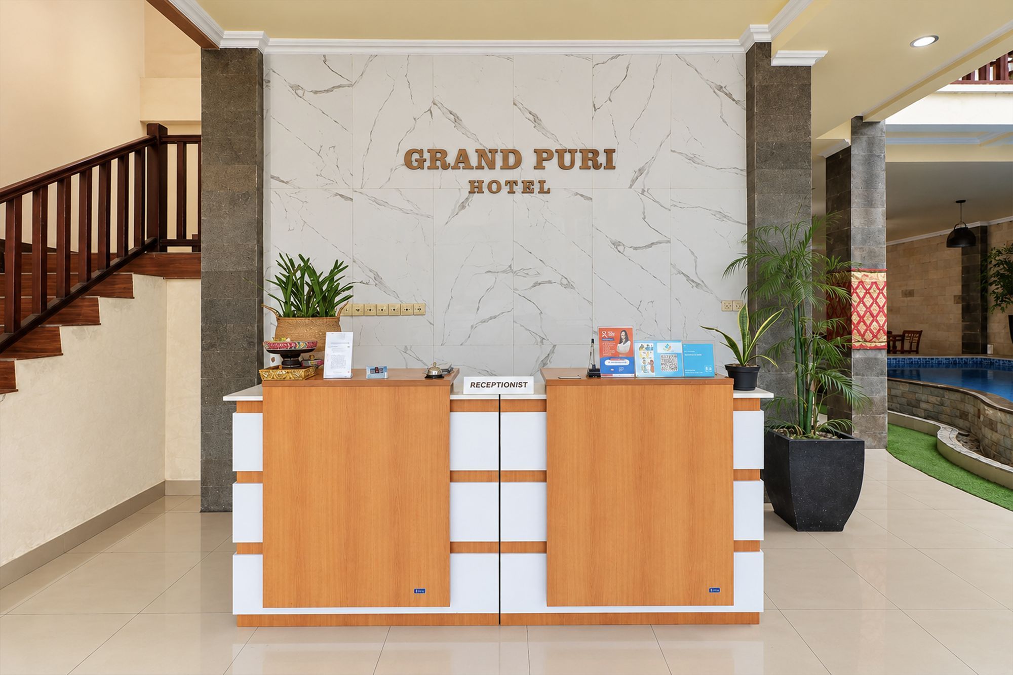 Mostrador de recepción en el Grand Puri Hotel, con un fondo de pared de mármol, una escalera a la izquierda y una zona de piscina visible al fondo.
