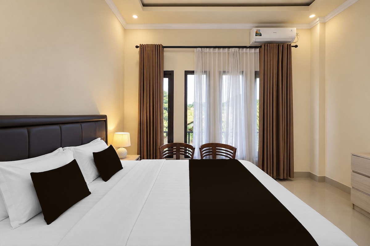 A hotel room with a bed, two chairs, a nightstand with a lamp, and a window with curtains.