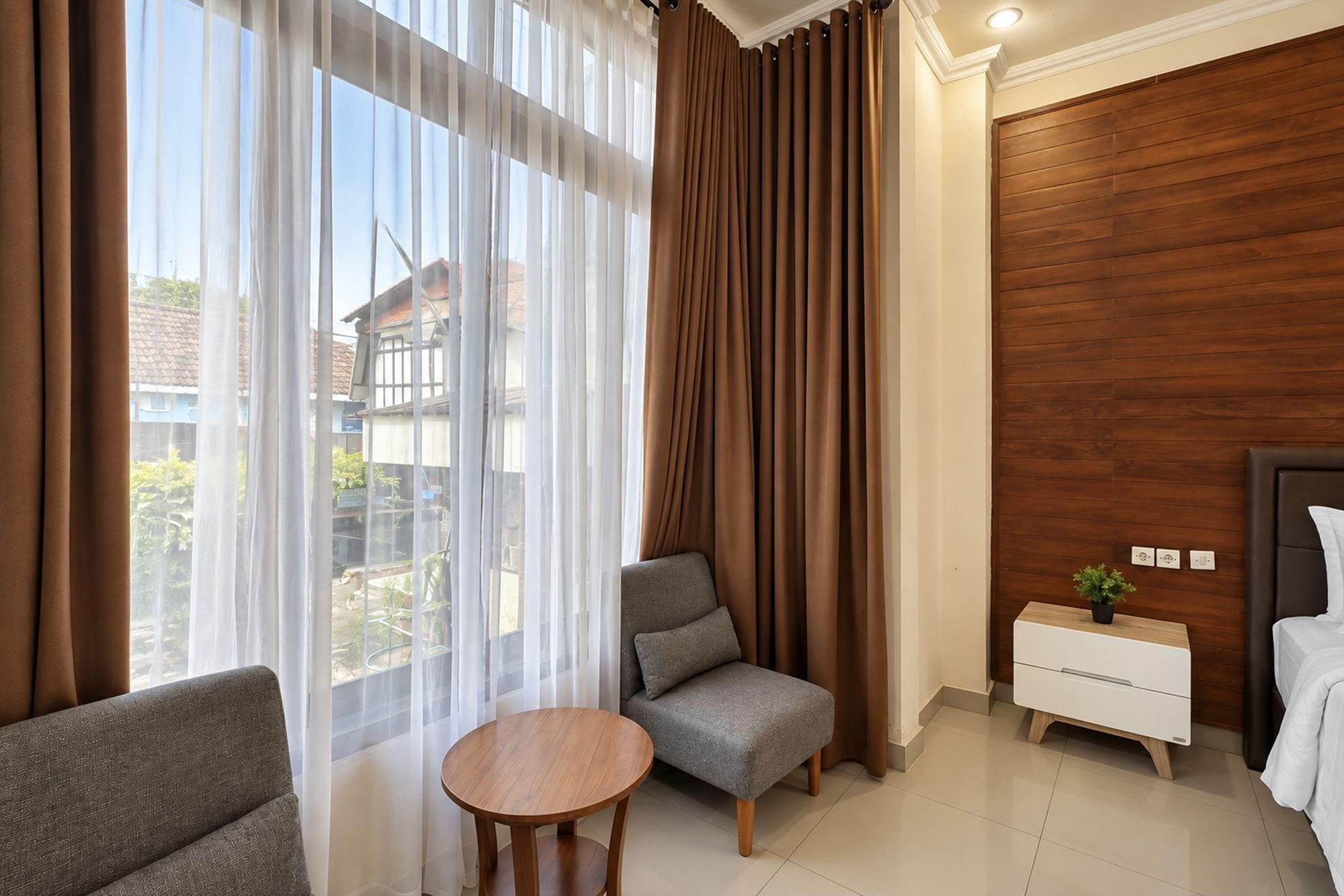 A hotel room with a bed, a small table, a chair, a nightstand, and a view of the outside.