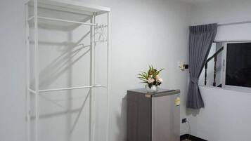 Una habitación con un armario metálico blanco, un refrigerador gris y una ventana con cortina.