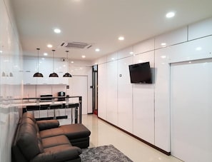 A modern living room with a sofa, a flat-screen TV mounted on the wall, and a kitchen area with a dining table and chairs.