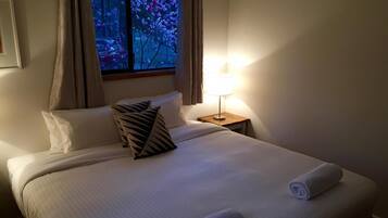 A hotel room with a bed, pillows, a nightstand with a lamp, and a window with curtains.