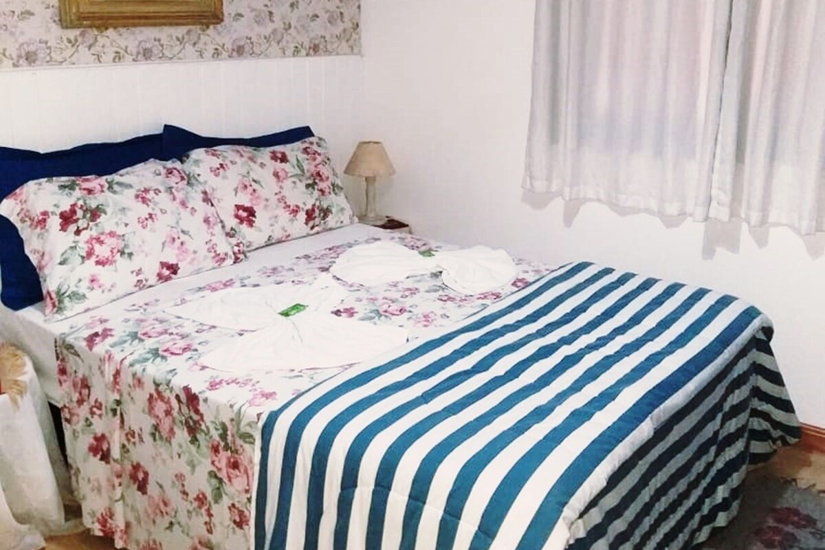 Un dormitorio con cama, mesita de noche, lámpara, un cuadro y ventana con cortinas.