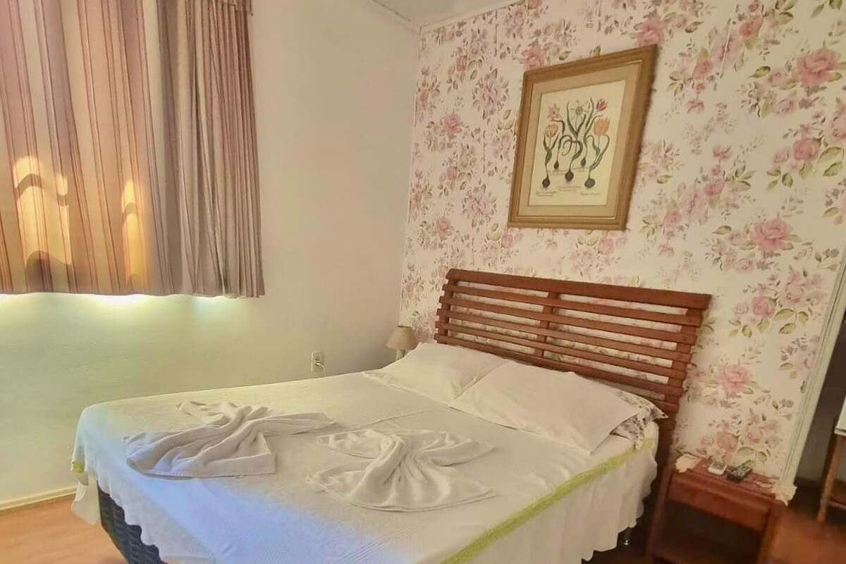 A neatly made bed with a wooden headboard, a floral wallpaper pattern on the wall, and a framed picture above the bed.