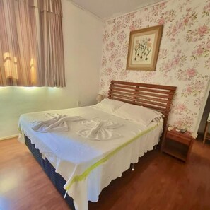 A neatly made bed with a wooden headboard, a floral wallpaper pattern on the wall, and a framed picture above the bed.