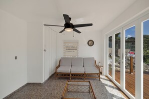 Een kamer met een plafondventilator, een bank, een glazen salontafel en uitzicht op het buitengebied.