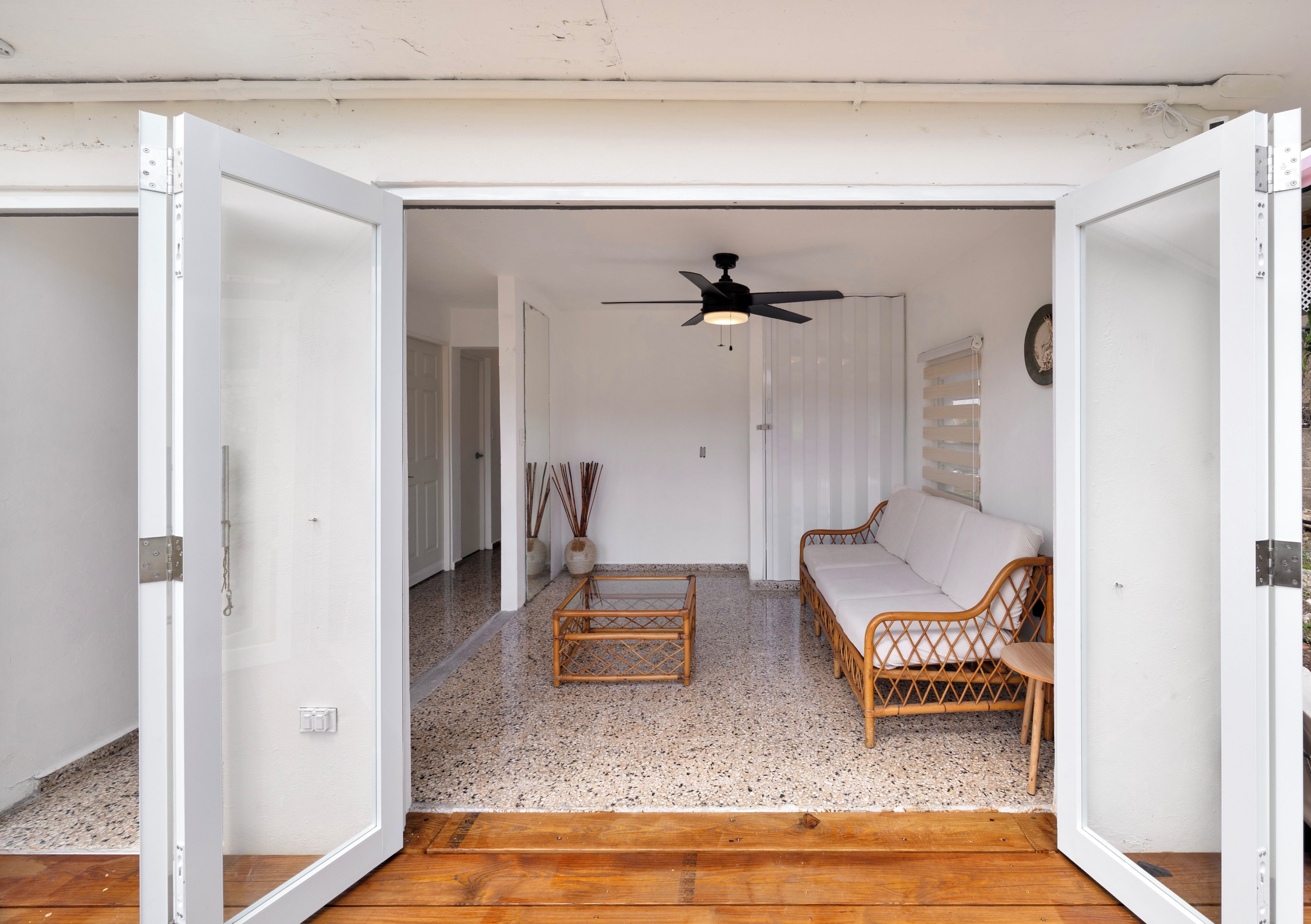 An open doorway leading to a living room with a ceiling fan, a sofa, a coffee table, and a wicker chair.