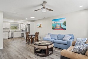 A modern kitchen with white cabinets, a dining area with a round table, and a living room with a blue sofa and a painting of a beach. - Shark Tooth Acres Manasota Key Retreat 4/3.5 (Englewood)