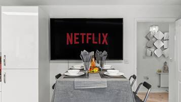 Ein Essbereich mit einem Tisch für zwei Personen, einem Fernseher, der 'NETFLIX' anzeigt, und einem dekorativen Wandbild.