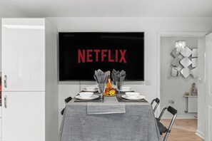 Ein Essbereich mit einem Tisch für zwei Personen, einem Fernseher, der 'NETFLIX' anzeigt, und einem dekorativen Wandbild.
