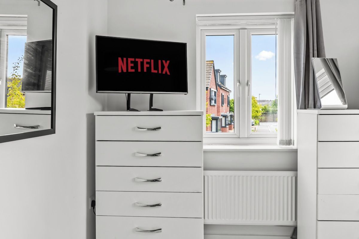 Ein modernes Schlafzimmer mit Fernseher, der 'NETFLIX' an der Wand anzeigt, einer weißen Kommode mit Schubladen und einem Fenster mit Blick auf Gebäude und Bäume.