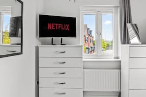Ein modernes Schlafzimmer mit Fernseher, der 'NETFLIX' an der Wand anzeigt, einer weißen Kommode mit Schubladen und einem Fenster mit Blick auf Gebäude und Bäume.