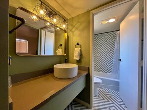 A bathroom with a round sink, a mirror with lights, a towel rack, and a patterned shower floor.