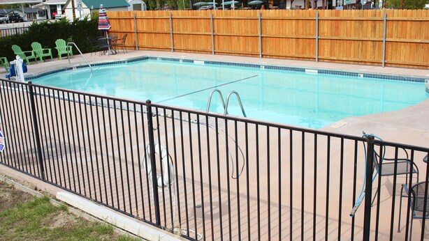 Una piscina con una cerca metálica, sillas verdes y una cerca de madera al fondo.