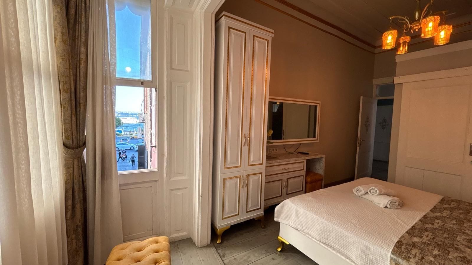 A hotel room with a bed, a wardrobe, a vanity with a mirror, and a window with a view of the street.