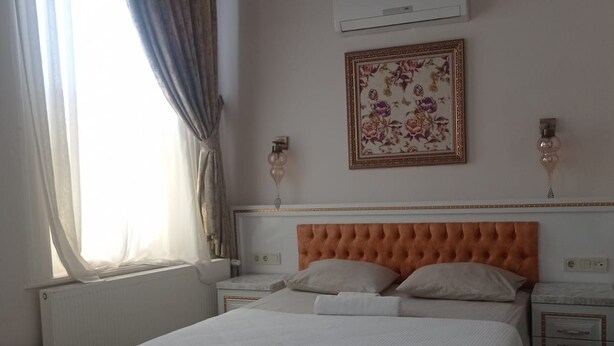 Un dormitorio con cama, una mesita de noche, aire acondicionado instalado en la pared, una obra de arte floral enmarco y una ventana con cortinas.
