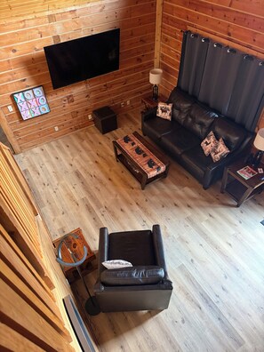 A log cabin interior with a flat-screen TV mounted on the wall, a leather sofa, a coffee table, and a small side table with a lamp.