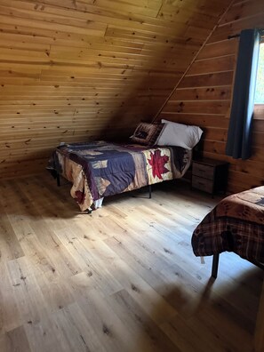 Ein Zimmer mit schrägen Wänden, Holzwänden, einem Bett mit gemustertem Überwurf, einem Nachttisch und einer Kommode.